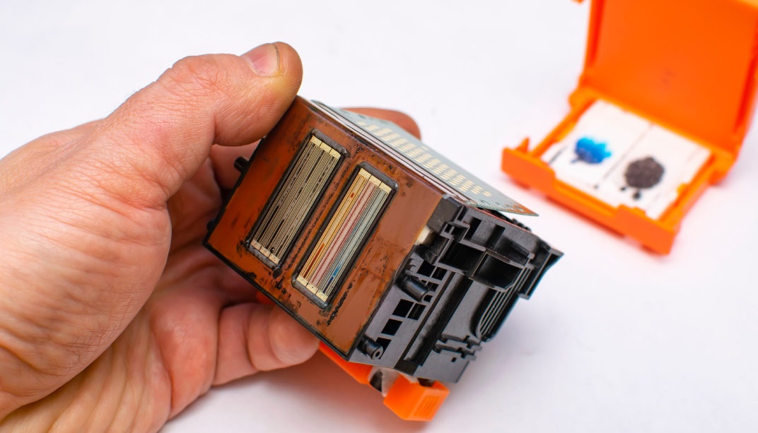 Druckkopf eines Tintenstrahldruckers mit eingetrockneten Tintenresten an den Düsen, daneben die orangefarbene Schutzkappe